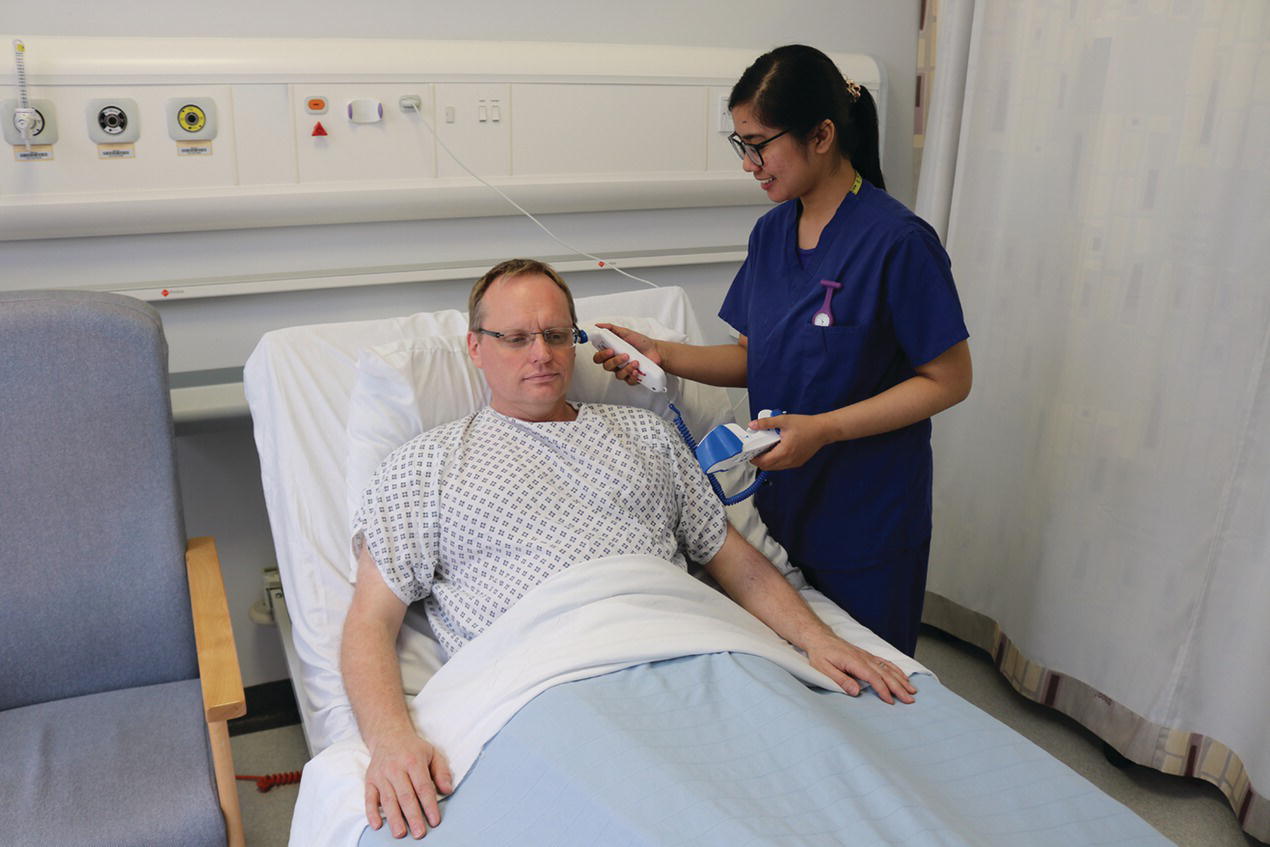 A woman standing beside a male patient (lying in bed) while inserting an electronic tympanic thermometer into the left ear of the patient.