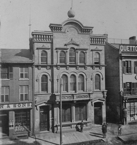 Old black and white photograph of a large three storey building with a storefront on the first floor. The building is flanked by other buildings with storefronts along King Street East.